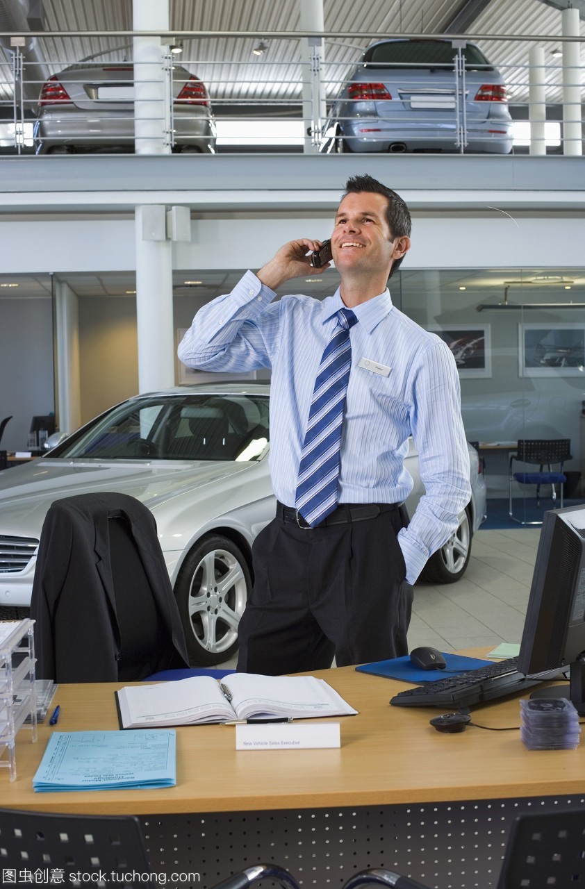 汽車銷售員站在柜臺(tái)旁的大型汽車陳列室里,用手機(jī)微笑著